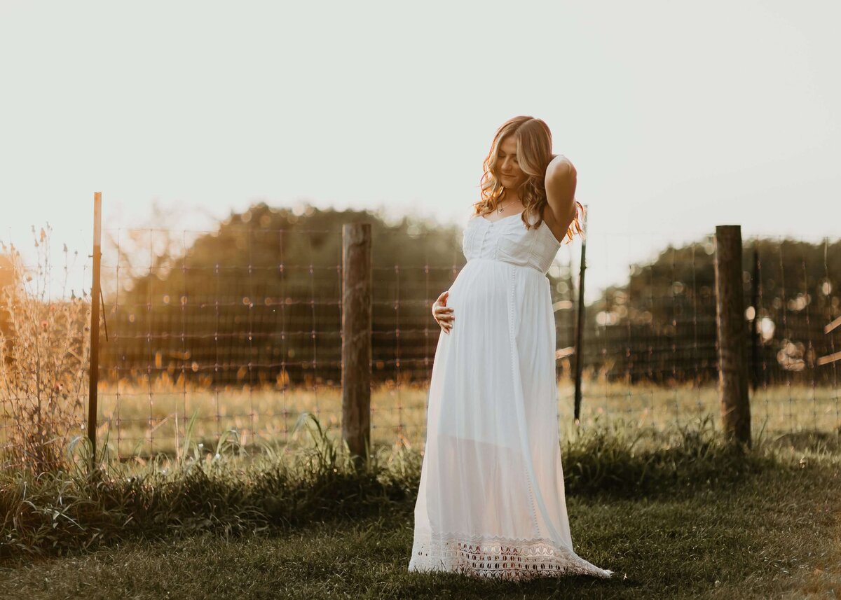 A pregnant woman in a white dress standing in a field at sunset is captured beautifully by a Pittsburgh maternity photographer.