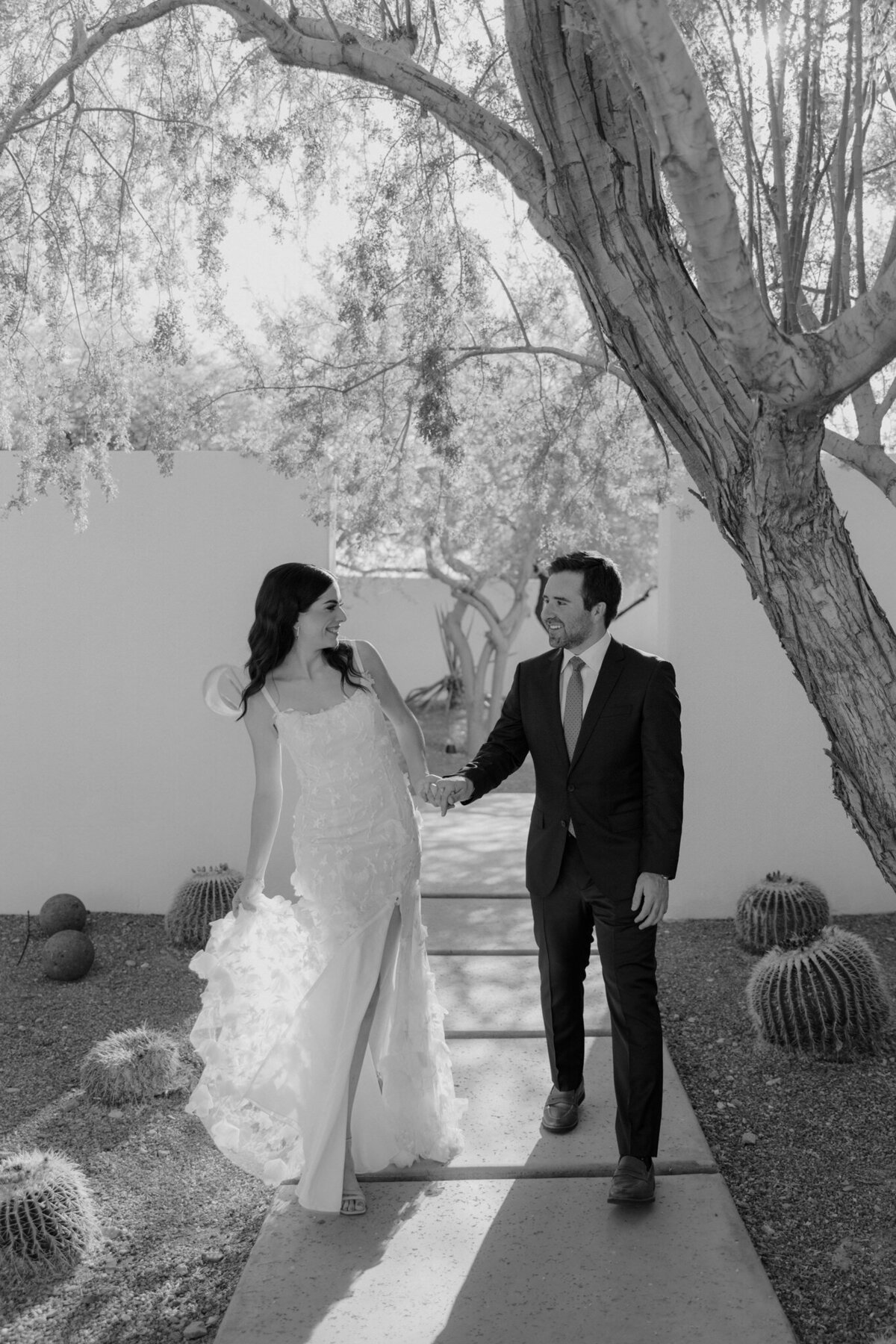 Bride wearing floral wedding dress and groom walking along paved path holding hands and smiling under a tree with cacti in the background.
