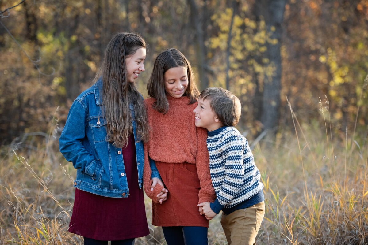 Three siblings giggle and hold hands while standing in a park trail at sunset for a Denver family photographer