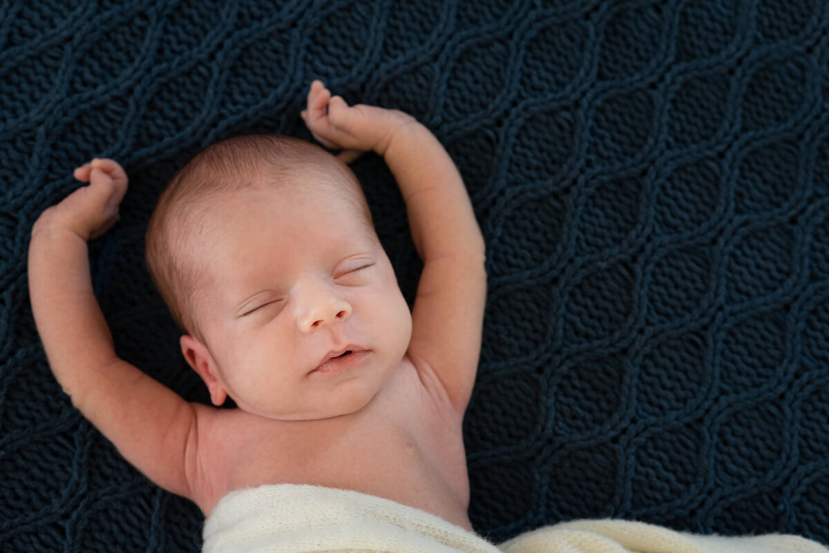 A newborn baby sleeping on a dark blue knit blanket stretches arms way up