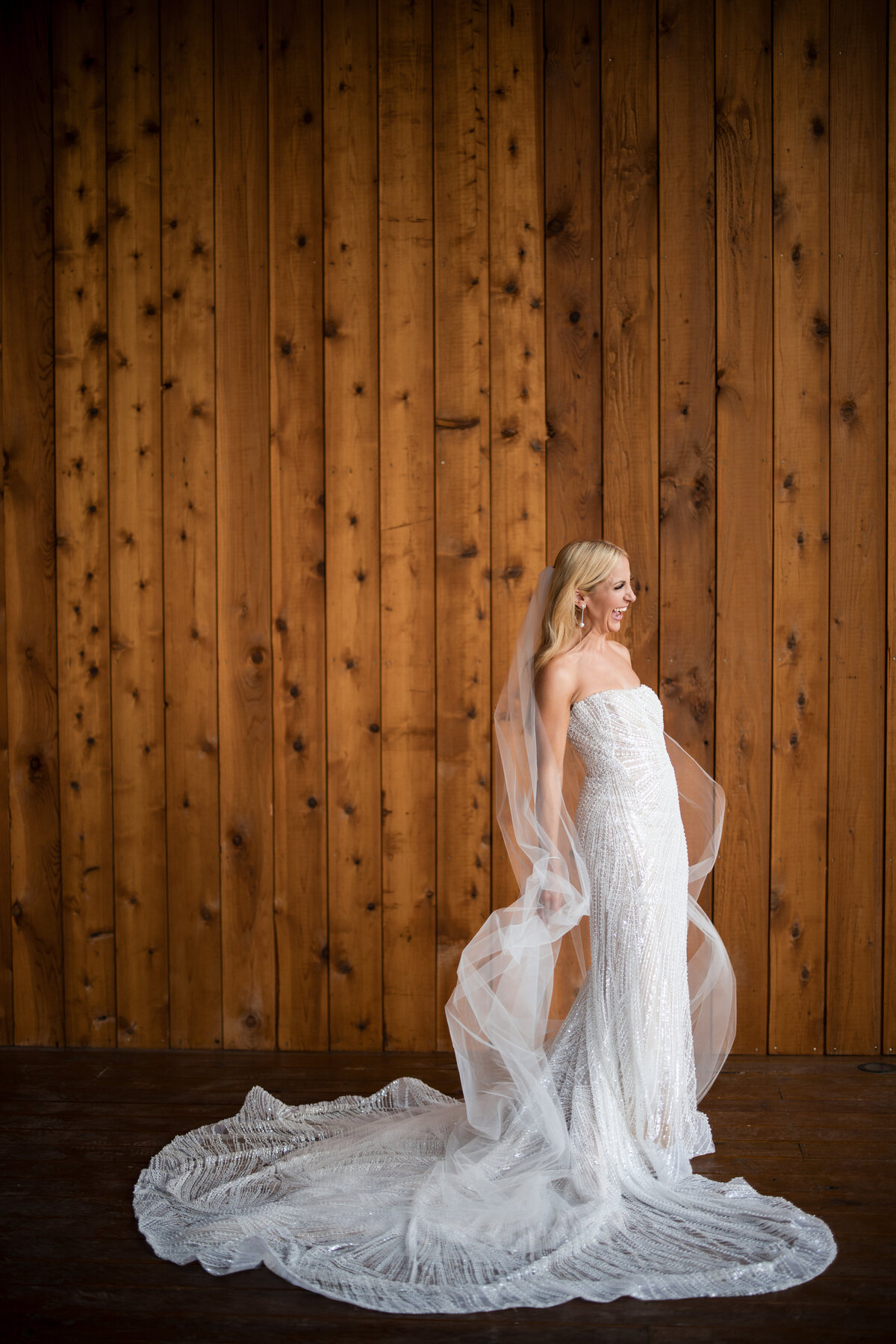 Bride in a white wedding gown stands against a wooden wall, with a long veil and train spread on the floor at a Southall destination wedding