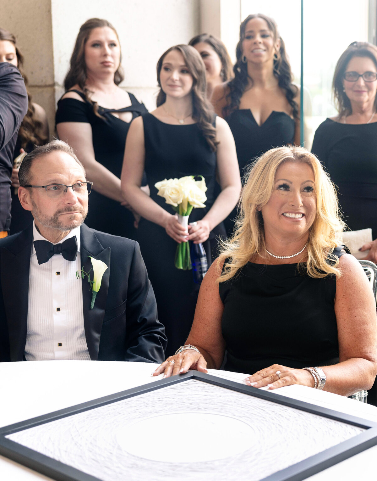 A group of people dressed in formal black attire stand and sit indoors. A man and woman are seated at a table in front of a large round artwork. The women in the background hold white flowers.