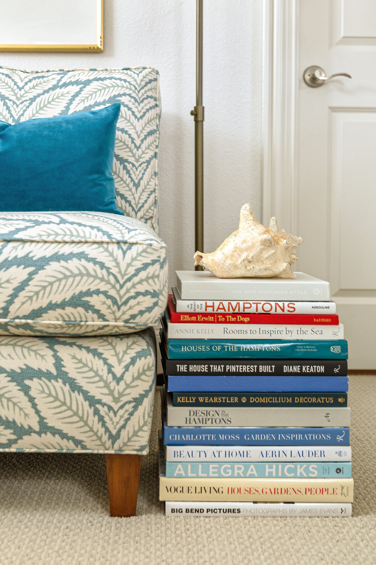Blue lounge chair with pattern fabric  and blue velvet accent pillow . Coffee table books stacked with seashell on top