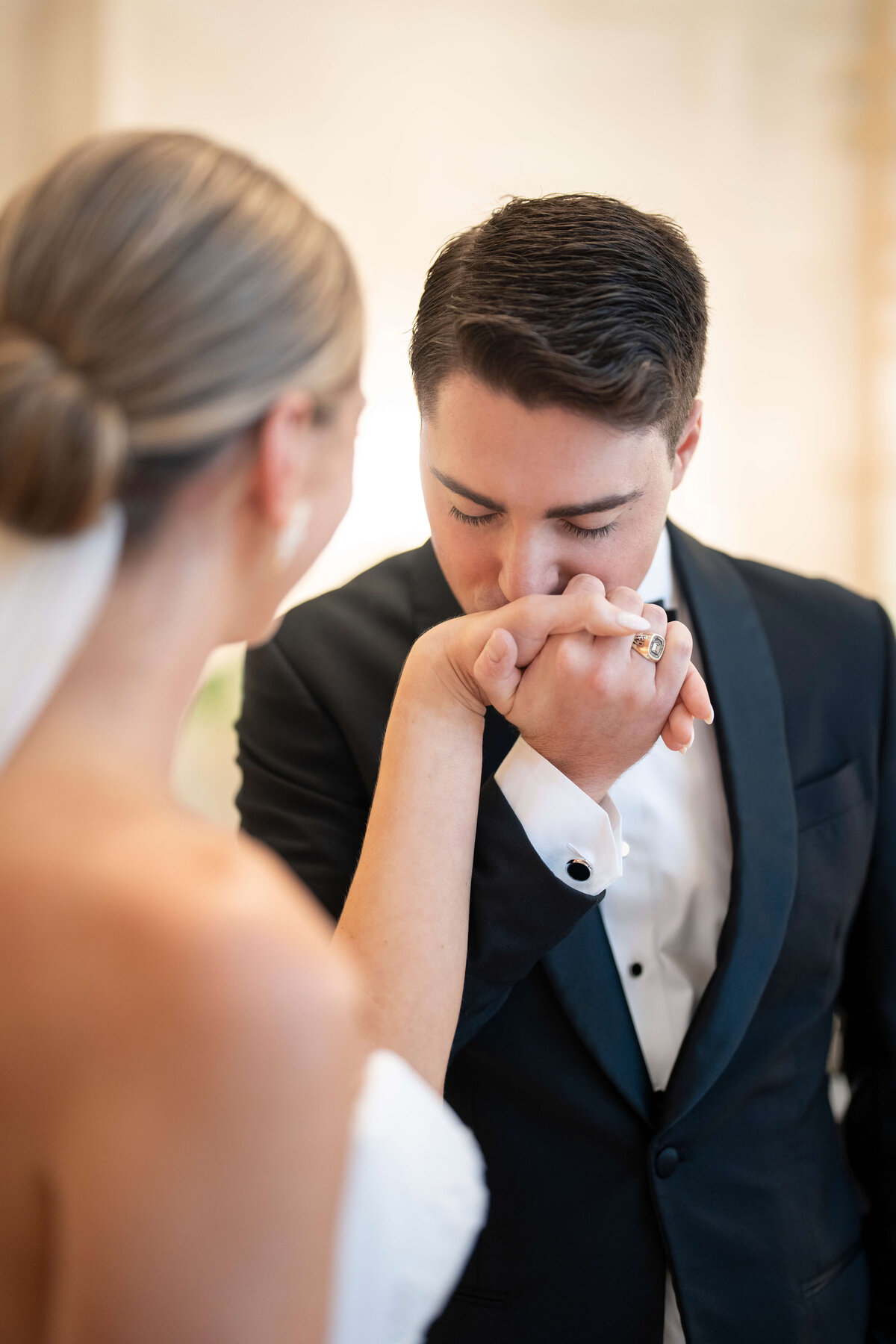 A groom in a tuxedo gently kisses the hand of his bride wearing a white wedding dress. The background is softly blurred, focusing on the intimate moment between them.