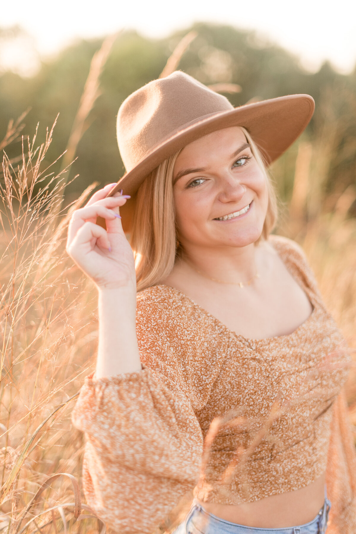 Senior girl smiling at camera with right hand on trendy hat brim on sunny day in Lancaster, Pennsylvania.