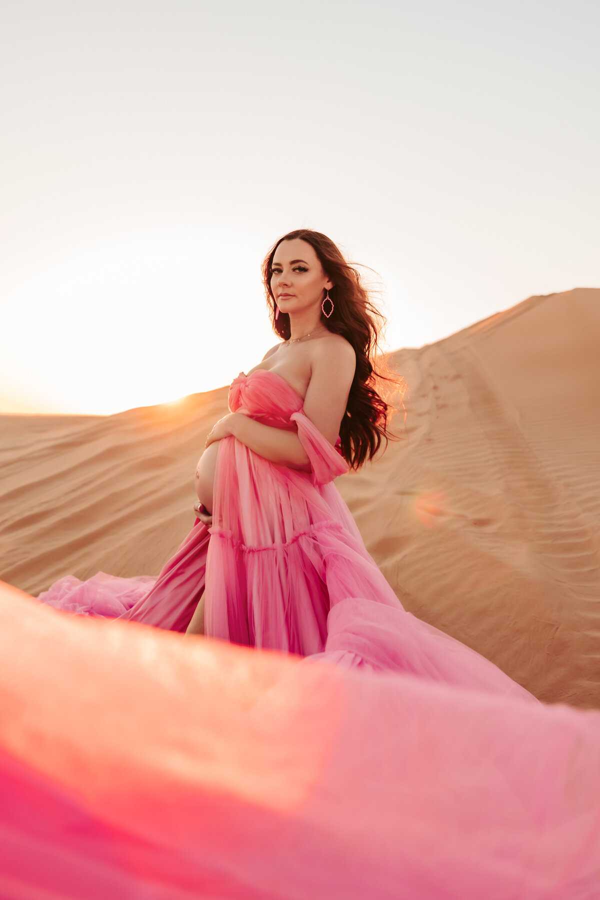 mother wearing bright pink tulle maternity gown holding her baby bump in the desert
