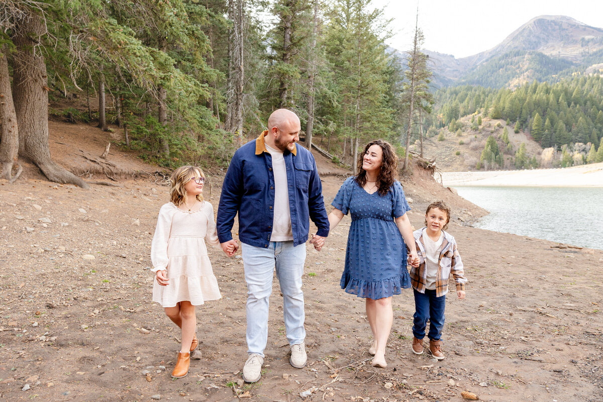 A family of four holds hands and walks barefoot along the shore of a mountain lake, with evergreen trees and a scenic view in the background.