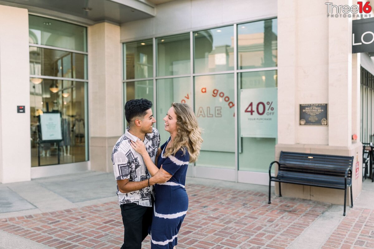 Engaged couple gaze at each other as they embrace in front of a shop in Downtown Brea