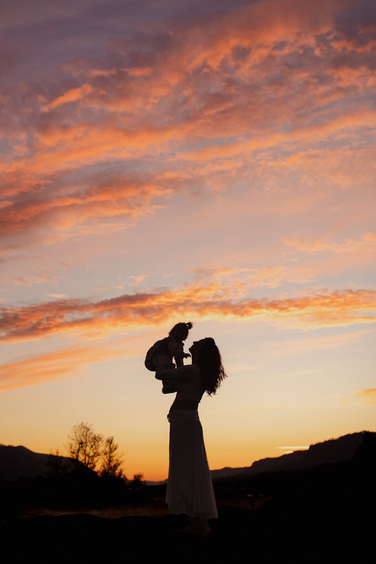Silhouette of mother holding her toddler up against a pink sunset. 