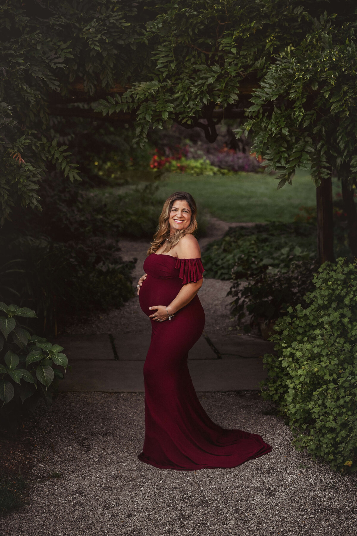 pregnany mom in a burgandy maternity gown standing under green  foliage arch in the summer at a park