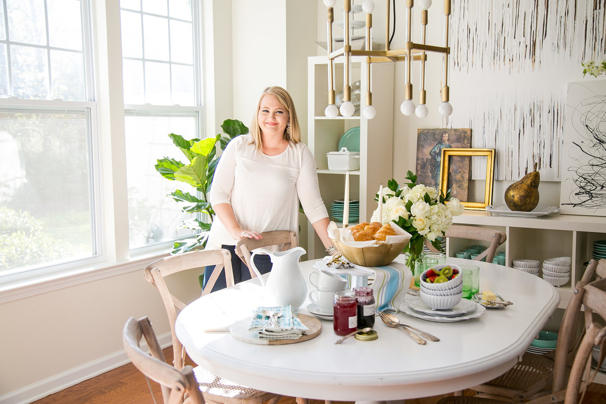 Blonde interior designer sets a dining table in beautiful sunlit dining room while smiling for brand photographs