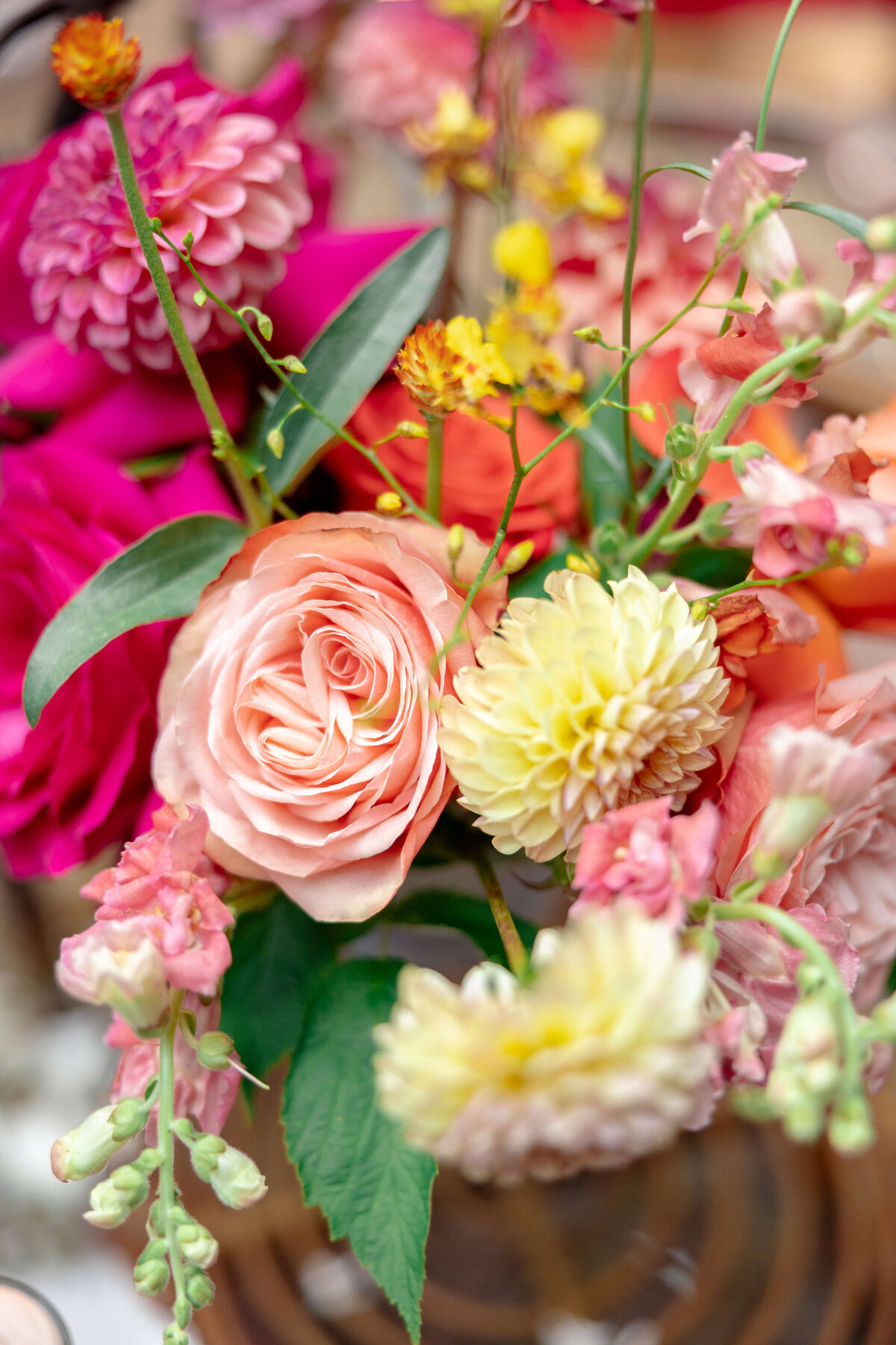 A vibrant bouquet of assorted flowers, including pink roses, yellow dahlias, and pink snapdragons, surrounded by green leaves. The flowers are arranged elegantly, showcasing a variety of colors and textures.