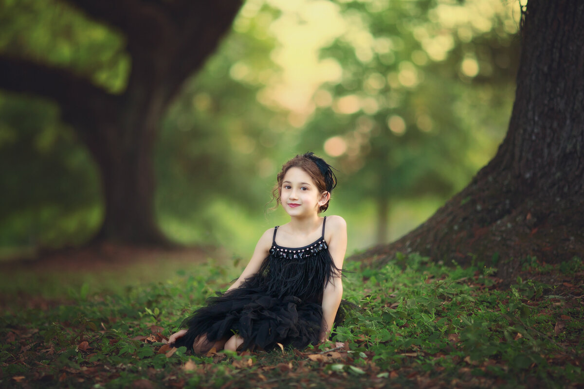 Young girl dressed in black tutu du monde with a feather headband.  She is sitting on the ground among the oak trees.  The sunlight is behind her.  She has dark hair in a bun and is looking towards the camera.