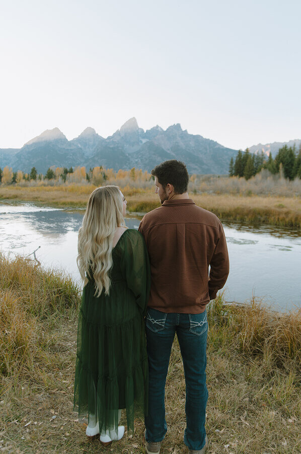 A newly engaged couple stand with their backs facing the camera. They're looking into each other's eyes with the Grand Tetons in front of them.