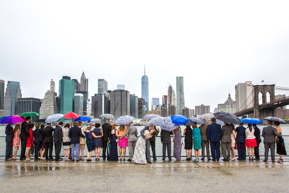 A bride and groom and their guests looking out over a city skyline while holding umbrellas.