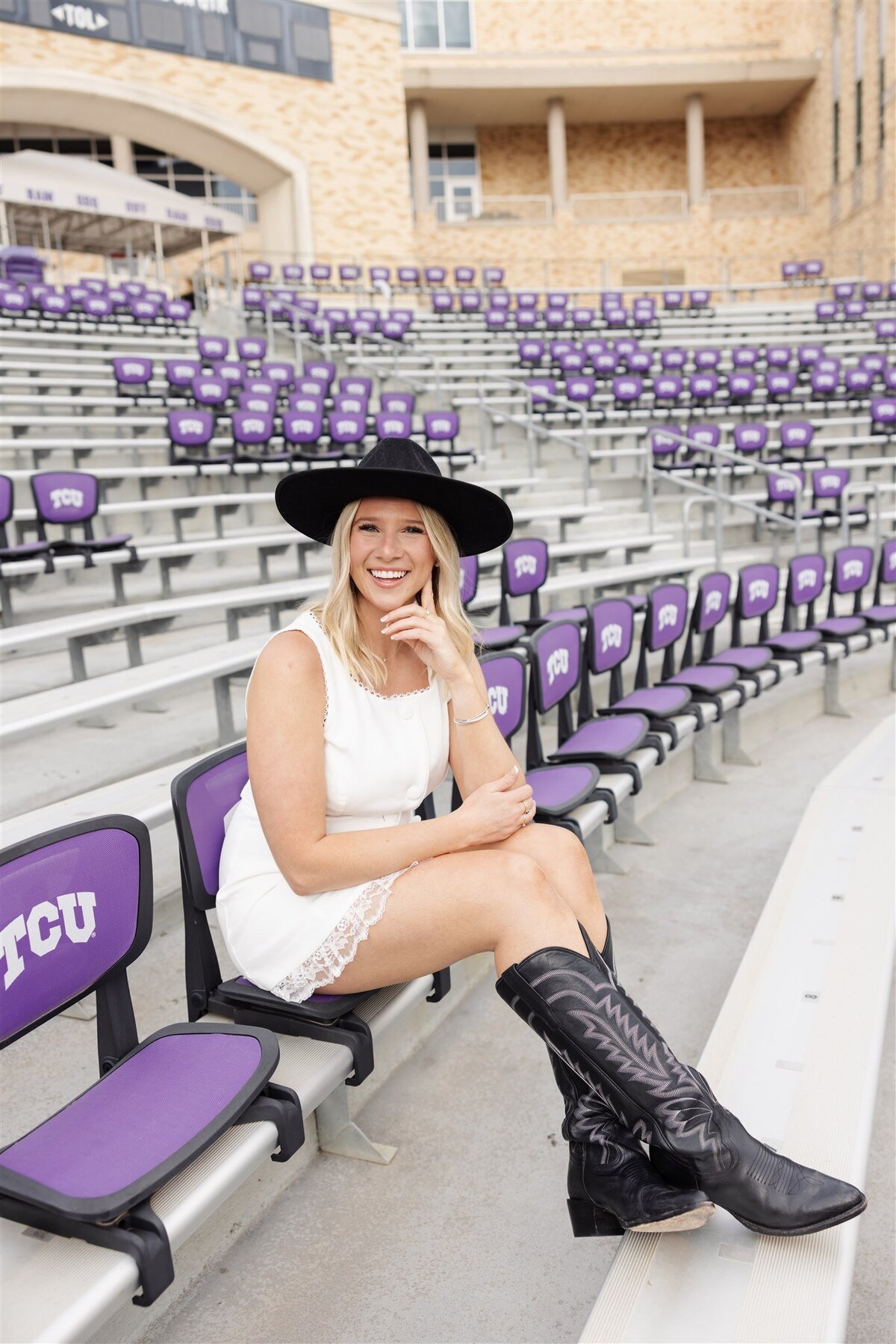 Senior girl poses on TCU stadium bleachers in white dress and black boots