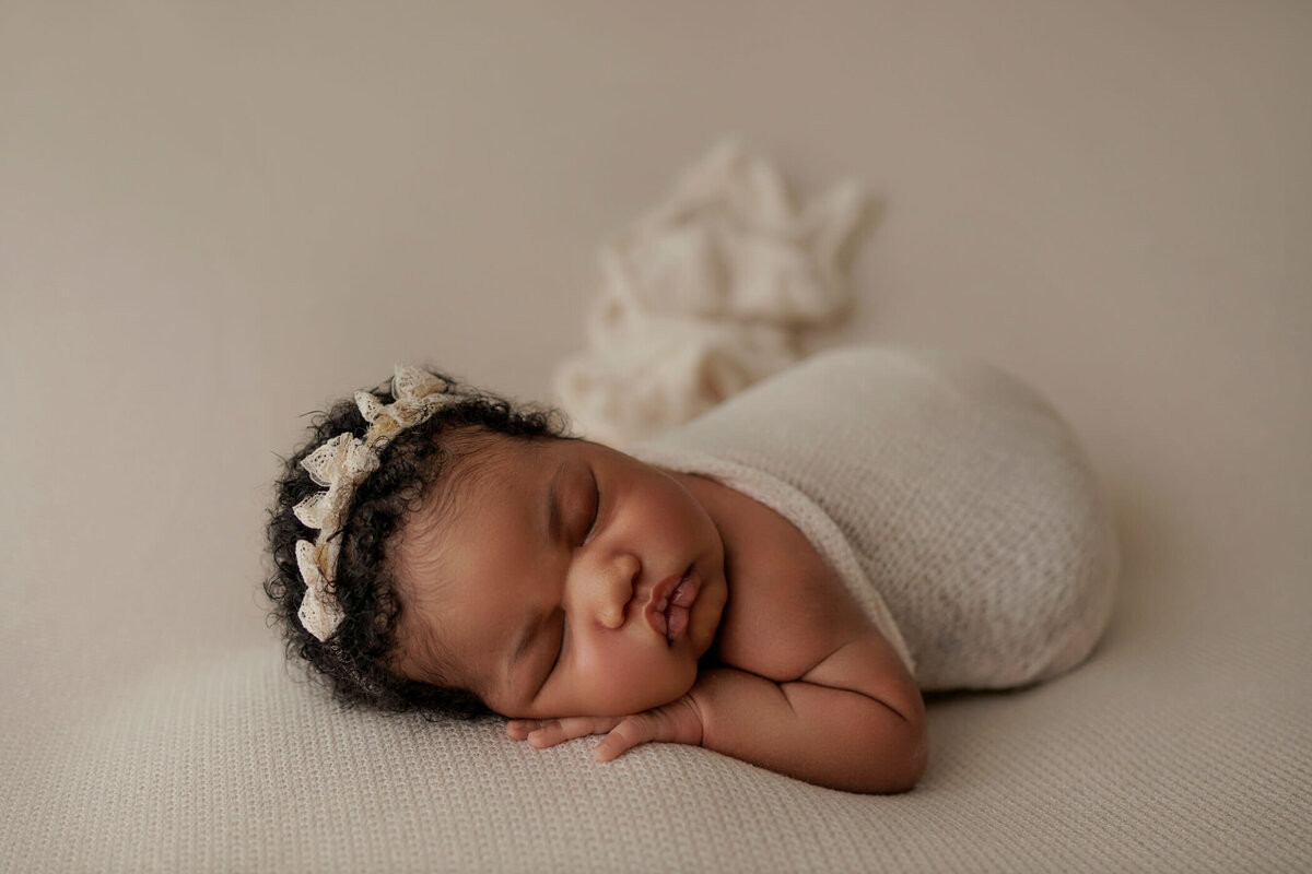 Newborn baby peacefully sleeping, swaddled in a light cream wrap with a matching headband adorned with small bows, against a neutral background.