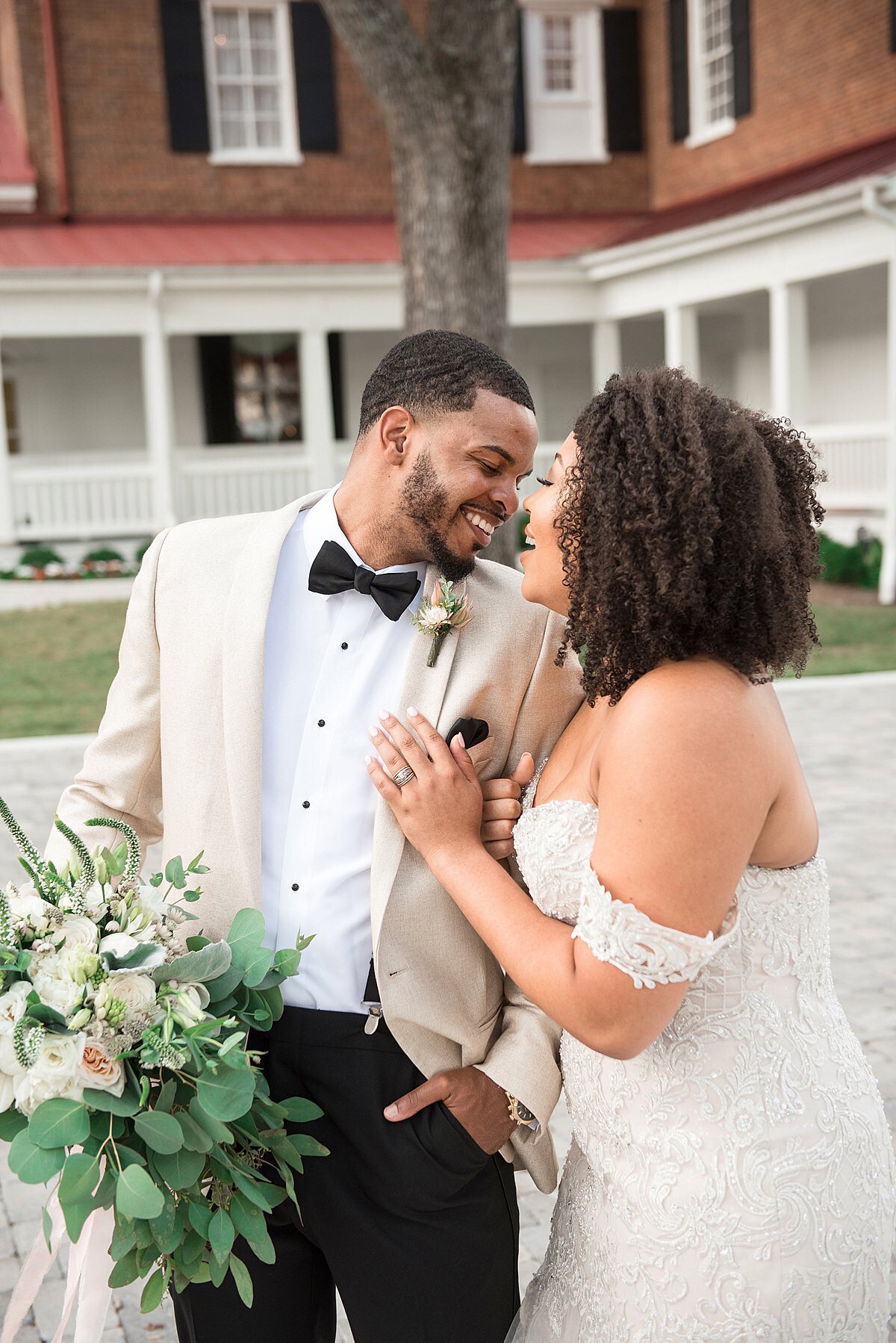 The groom, Alex Sweet, wearing a black and white tuxedo holding the bridal bouquet of greenery, white, ivory and blush flowers touches noses with his wife, Jasmine Sweet who is wearing an off the shoulder lace mermaid dress while standing outside in the courtyard behind Ravenswood Mansion