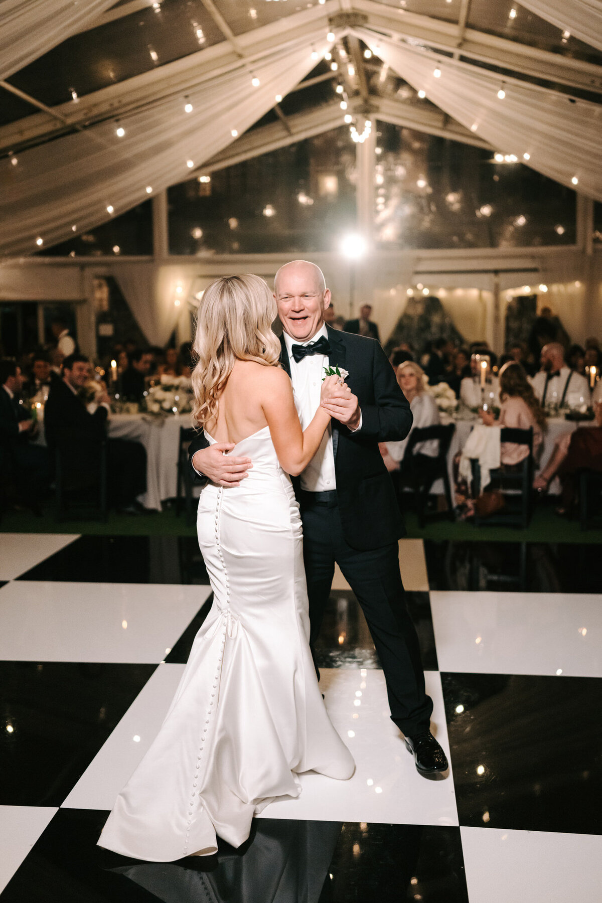 A photograph in color of Claire and her father on her wedding day at Woodbine Mansion in Austin, Texas. The bride and her father are alone on the black and white checkered dance floor. They are in a tent draped in white fabric with cafe lights lighting the room. The father of the bride is facing the camera with a huge smile on his face. It is a full length photo showing the bride’s mermaid style, low back, sleeveless dress and the father of the bride in a black and white tuxedo. Wedding photography by Stacie McChesney/Vitae Weddings.