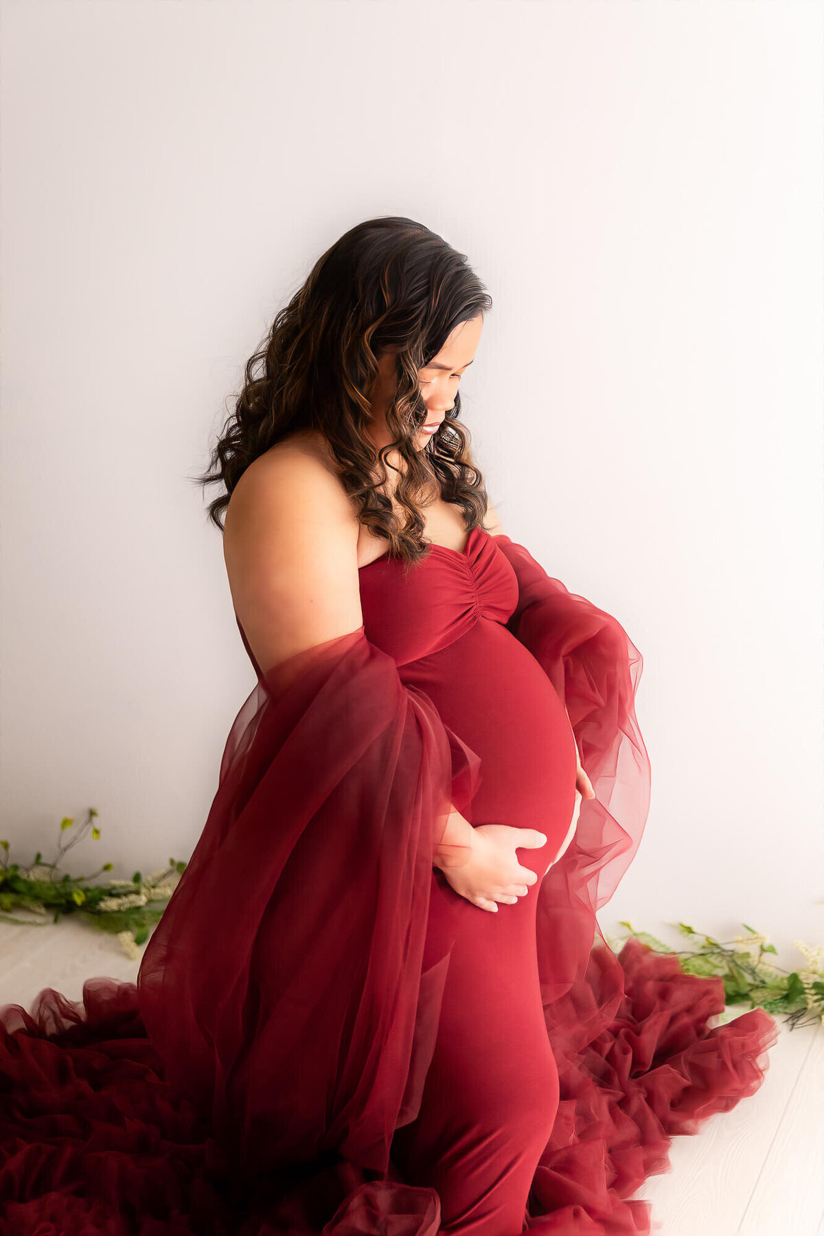 A pregnant woman in a stunning red gown poses gracefully for a photo, showcasing her beautiful baby bump.