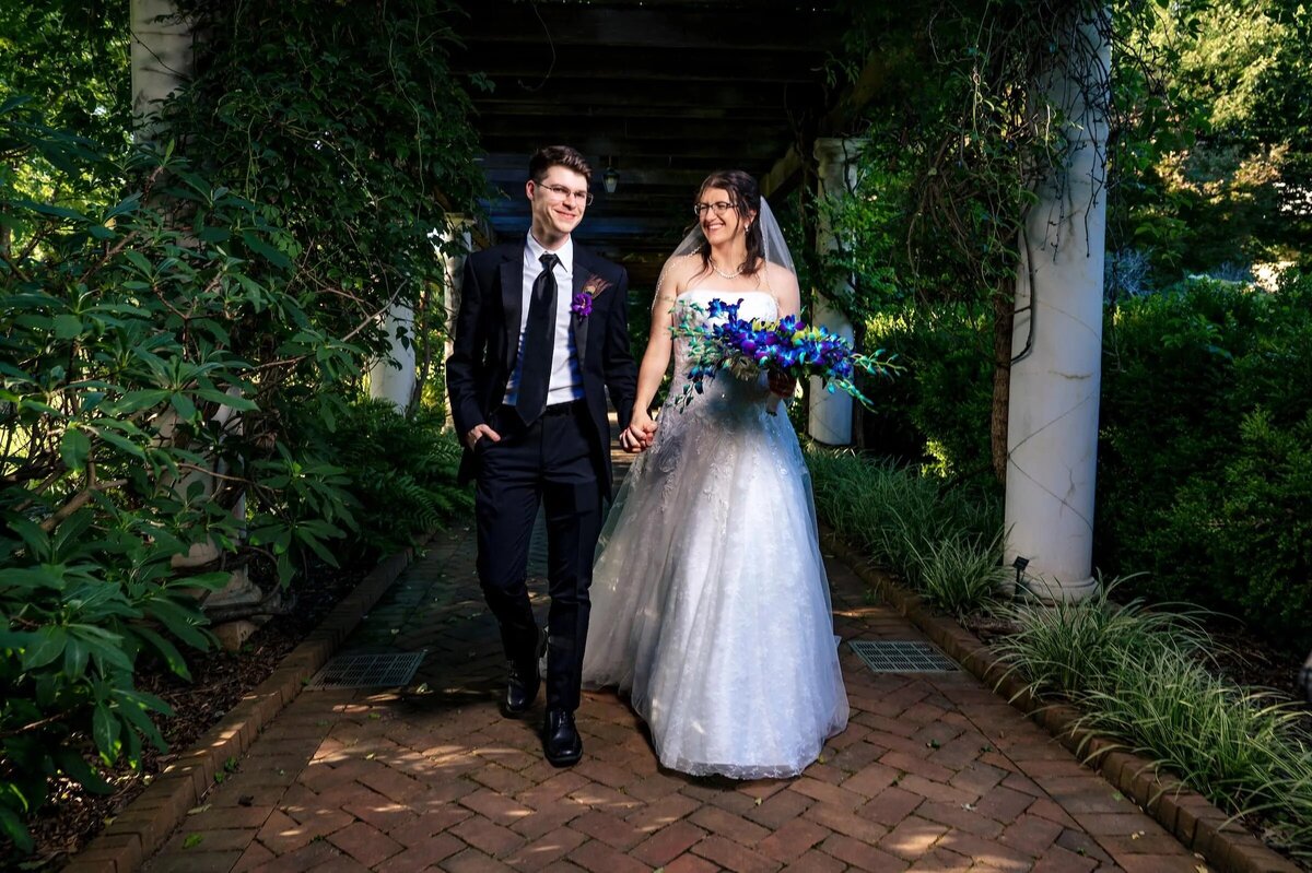 A smiling bride in a white gown with a blue bouquet and groom in a black suit walking hand-in-hand down a garden pathway.