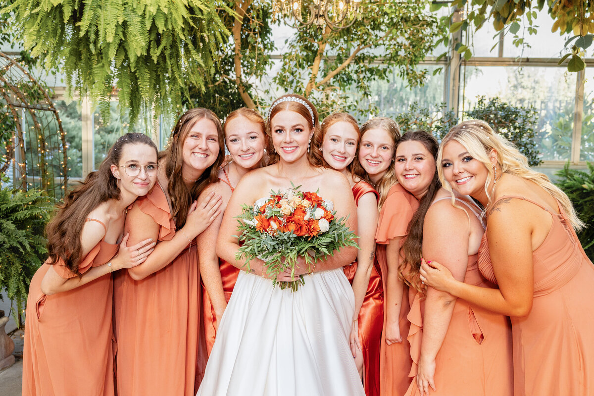 The bride poses with her bridesmaids, all in burnt orange dresses, under vibrant hanging greenery, holding her bouquet of orange and white flowers.