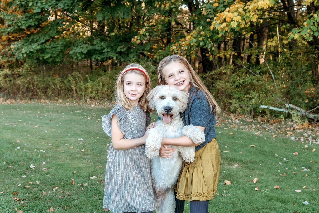 Two sisters hugging their fluffy white dog during Connecticut family photographer fall portrait session in park setting