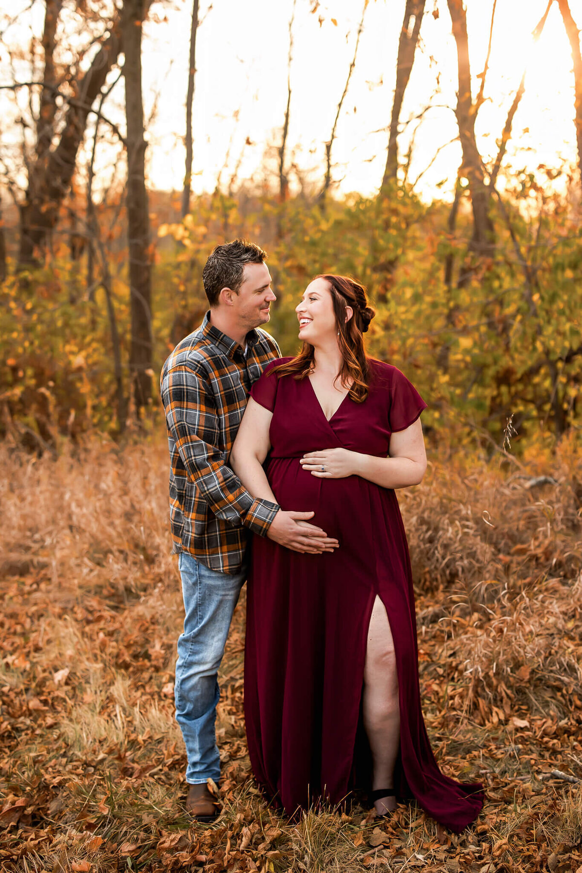 A couple stands outdoors in autumn; the woman, wearing a burgundy dress and visibly pregnant, smiles at the man. Captured by a Bloomington maternity photographer, they embrace among trees and fallen leaves in this beautiful pregnancy photography moment.