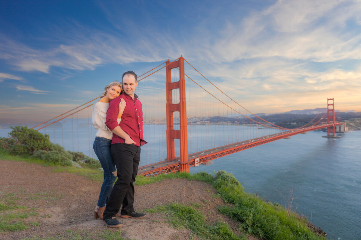 Engaged woman holds man from back cuddling close to him.  He looks back at her while she looks at the camera. They stand on a ledge overlooking the golden gate bridge and san francisco bay area. Photo by wedding photographer, sacramento, ca philippe studio pro.
