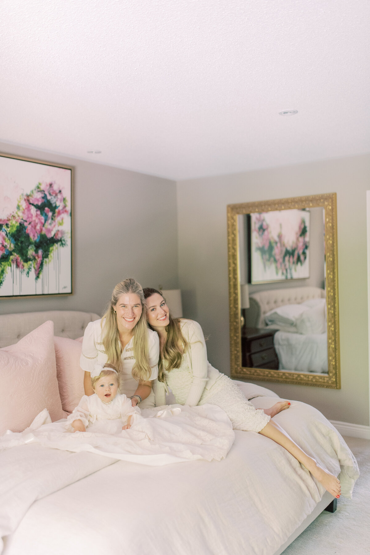 Serene portrait of mother and godmother cradling baby on soft bedding, mirror reflecting floral tones.
