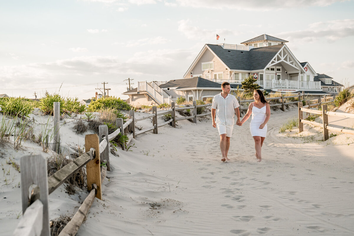 A couple holding hands and walking barefoot on a sandy beach path near a large house with a coastal design.