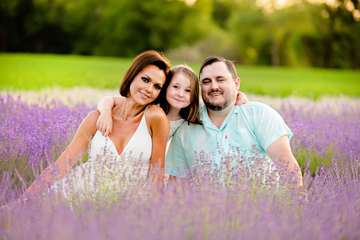 mom and dad with their daughter sitting with lavender all around them for their Burlington, On lavender family photographer session
