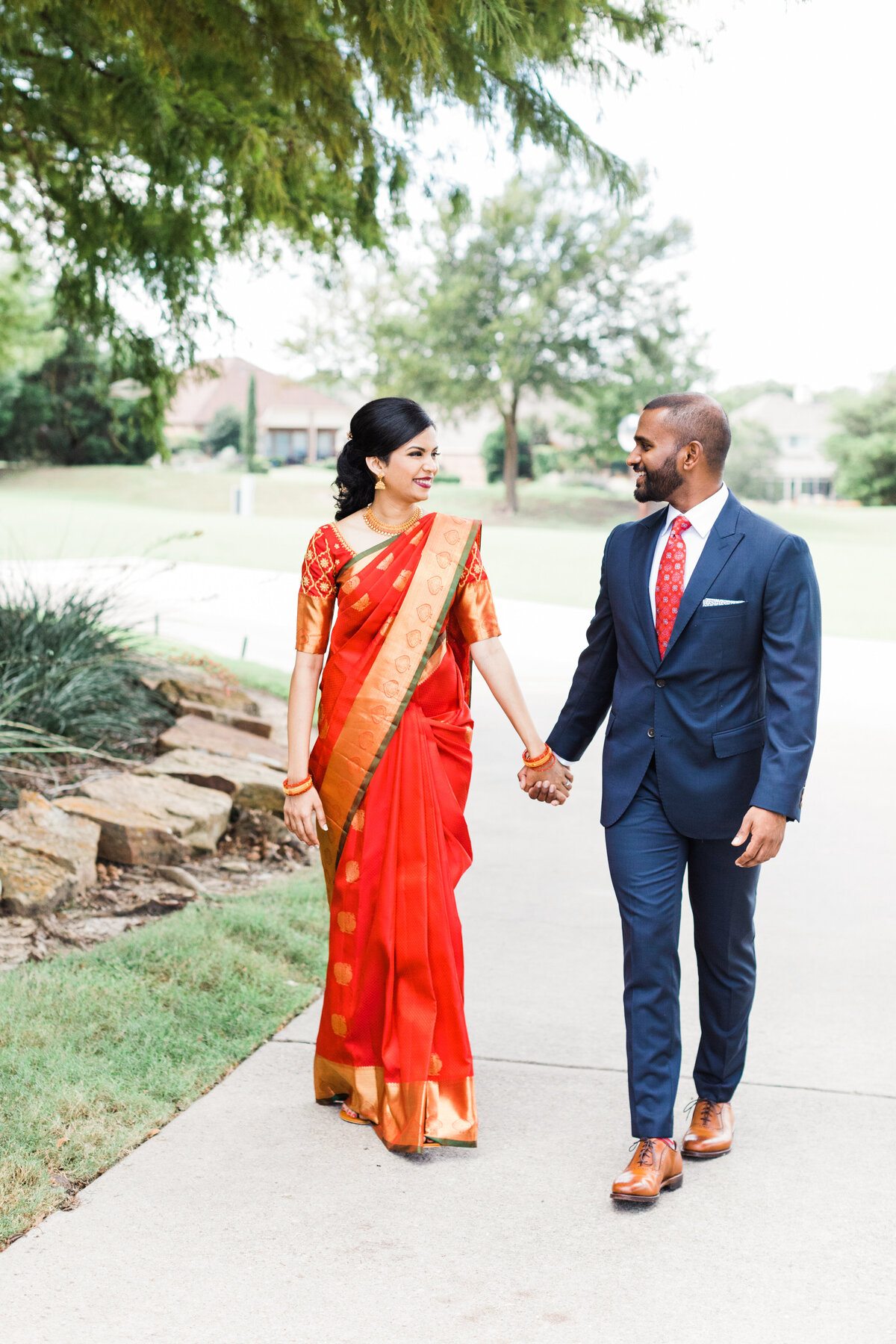 An Indian couple holding hands and looking into each other's eyes while walking down the road during their engagement photoshoot in DFW, Texas. The woman on the right is wearing traditional Indian attire while the man on the right is wearing a navy suit with a red tie and pocket square.
