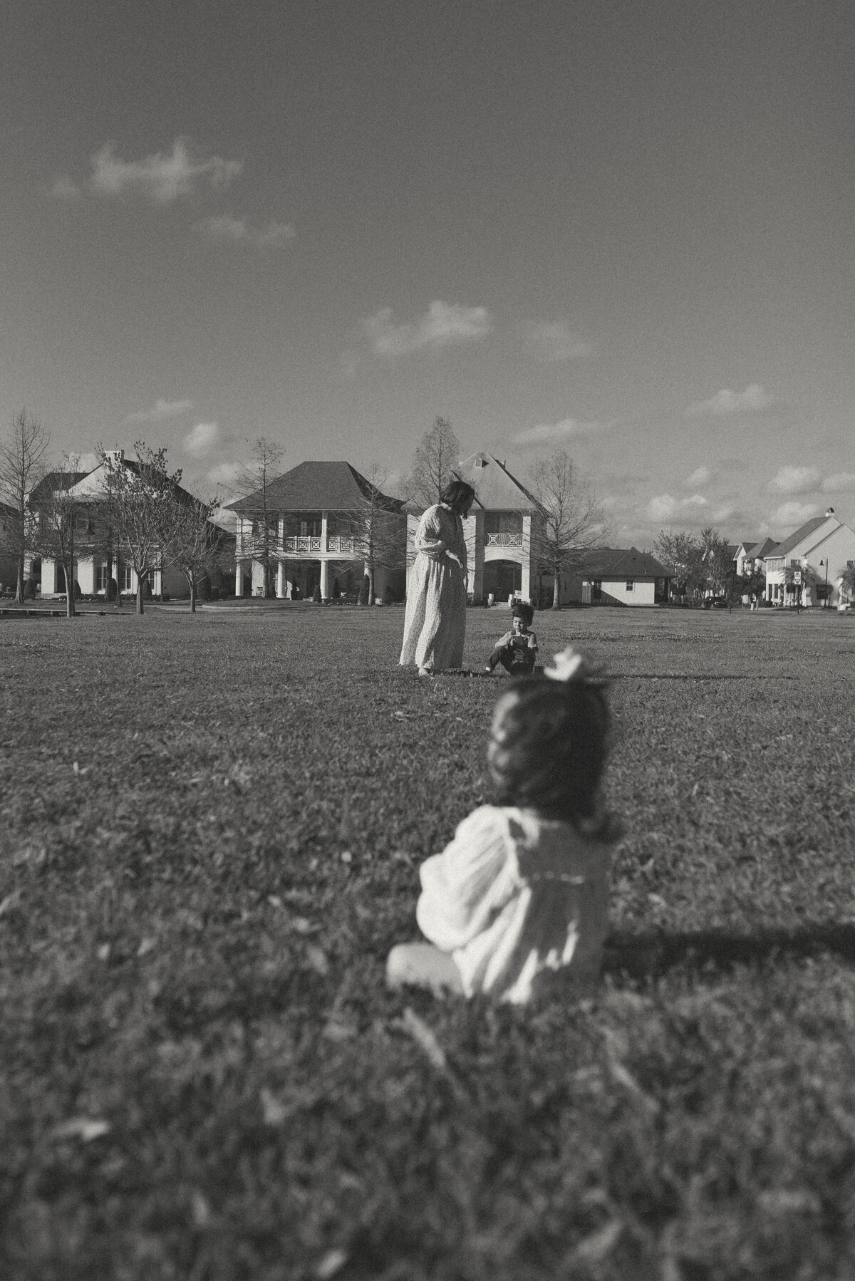 black and white photo of a toddler girl sitting in some grass with her mother and brother in the background in the distance.