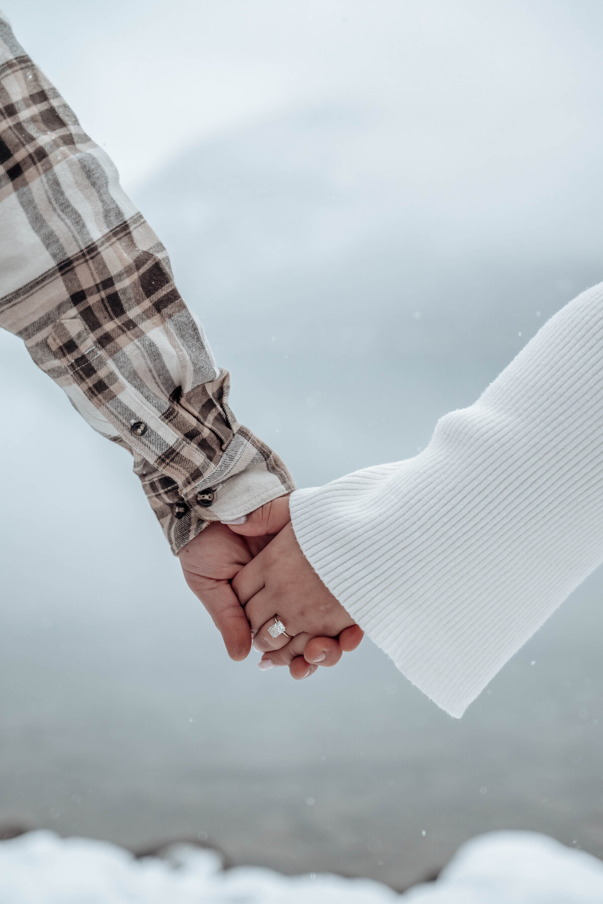 Engaged couple holds hands in snowy field with ring detail in focus.