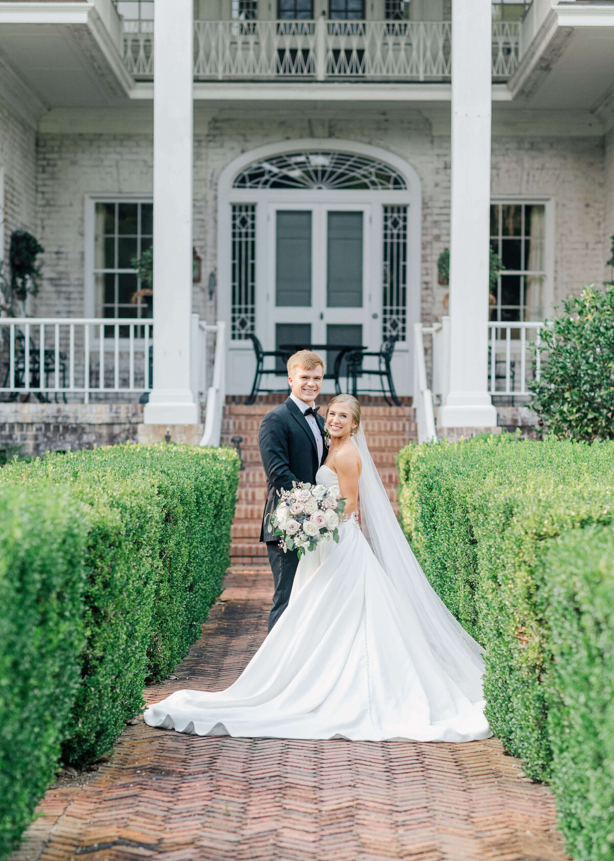 Alabama wedding couple standing inf ront of a white building surrounded by green hedges smiling at camera