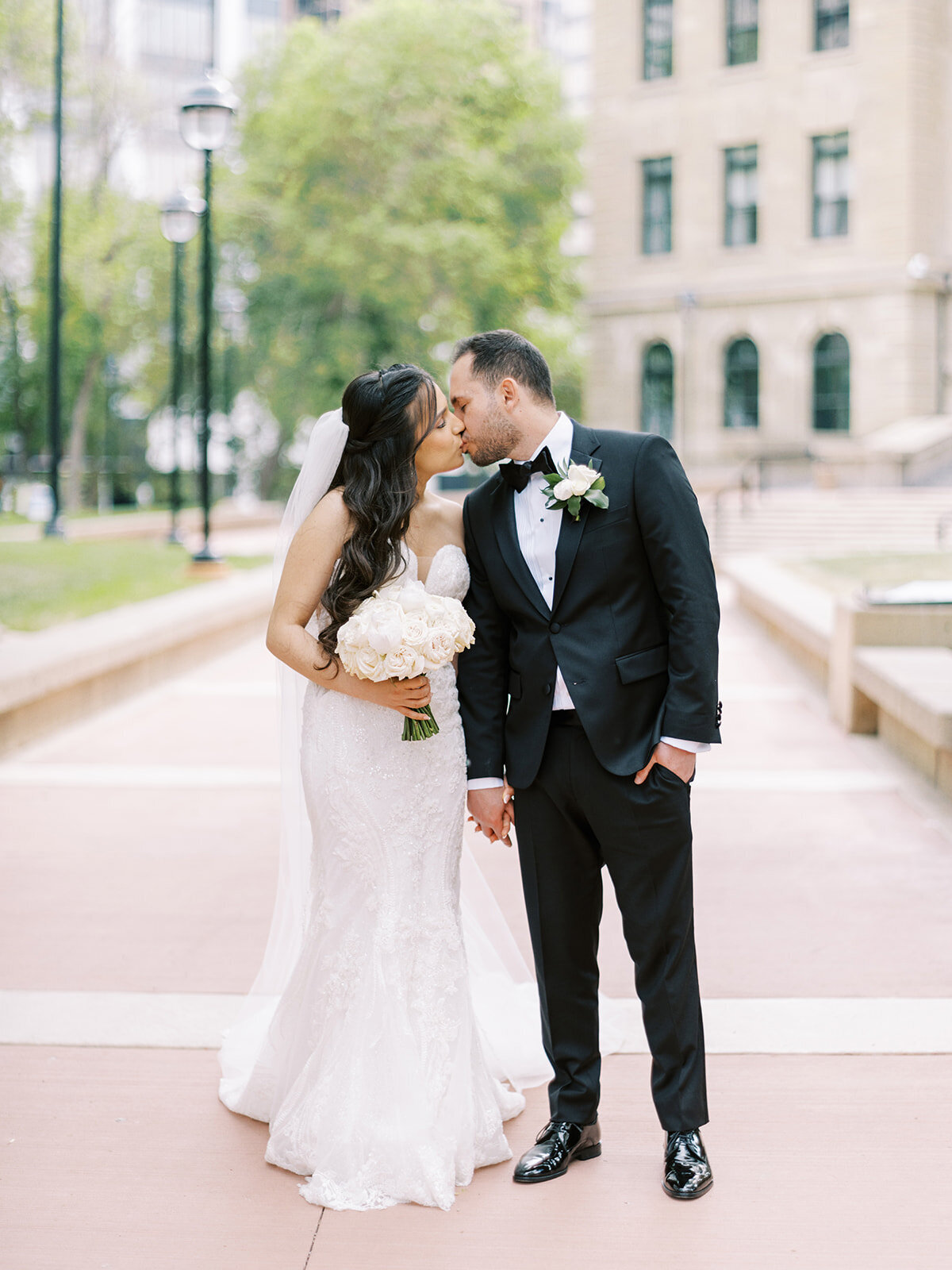 A bride in a white dress and veil holding a bouquet kisses a groom in a black suit and bow tie outdoors on a sunny day, capturing the essence of a classic Calgary wedding.