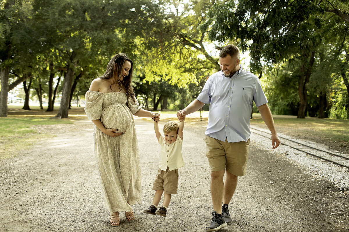 A joyful maternity photo at Trinity Park, with mom, dad, and big brother walking hand-in-hand along a scenic path, smiling and enjoying the moment as a family, captured by Chunky Monkey Photography.