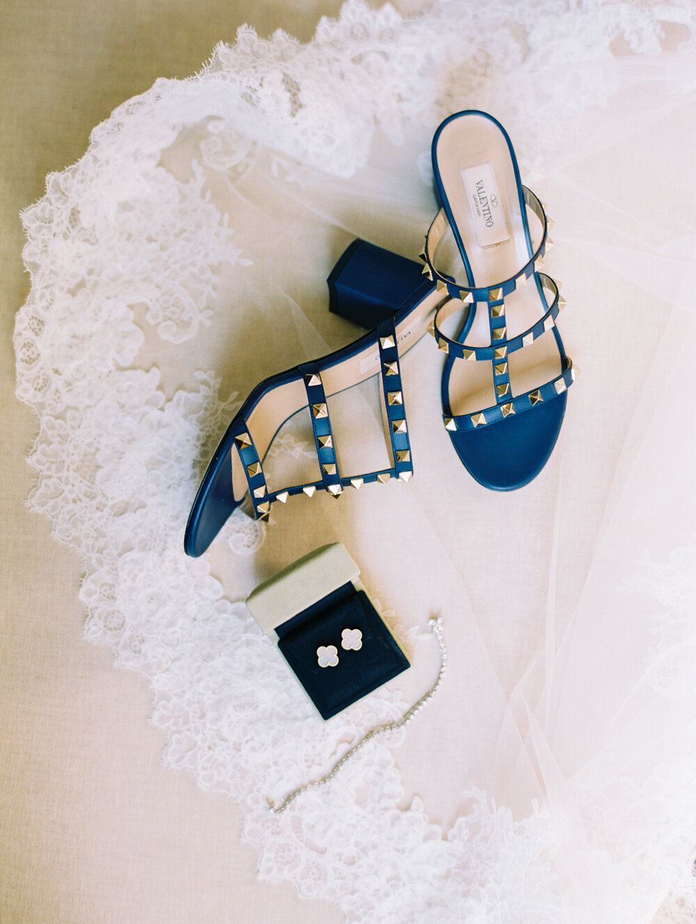 The photo was taken at The Hamptons, New York, a stunning waterfront estate tented wedding captured by photographer Bonnie Sen. In the scene, a pair of blue strappy high-heeled sandals with gold studs is elegantly placed on a white lace fabric, adding a touch of glamour to the setting. Adjacent to the sandals sits an open navy blue jewelry box, revealing a pair of exquisite white floral earrings nestled inside. Completing the ensemble, a delicate pearl bracelet lies gracefully beside the jewelry box, enhancing the overall elegance of the accessories.