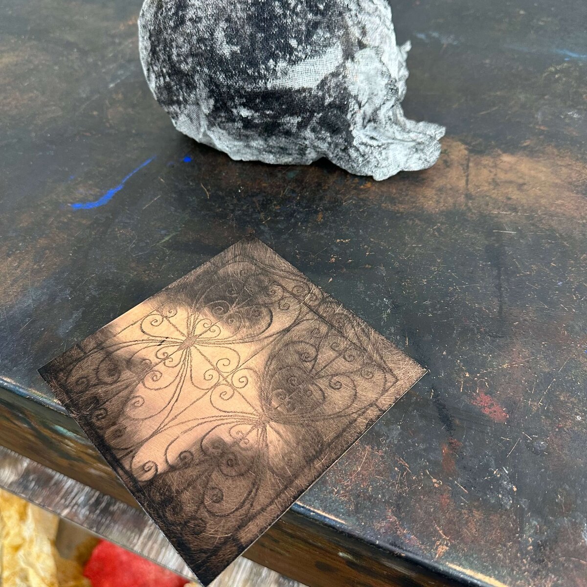 A copper etching plate with a heart-inspired scroll design sits inked on a dark tabletop, ready for printing.