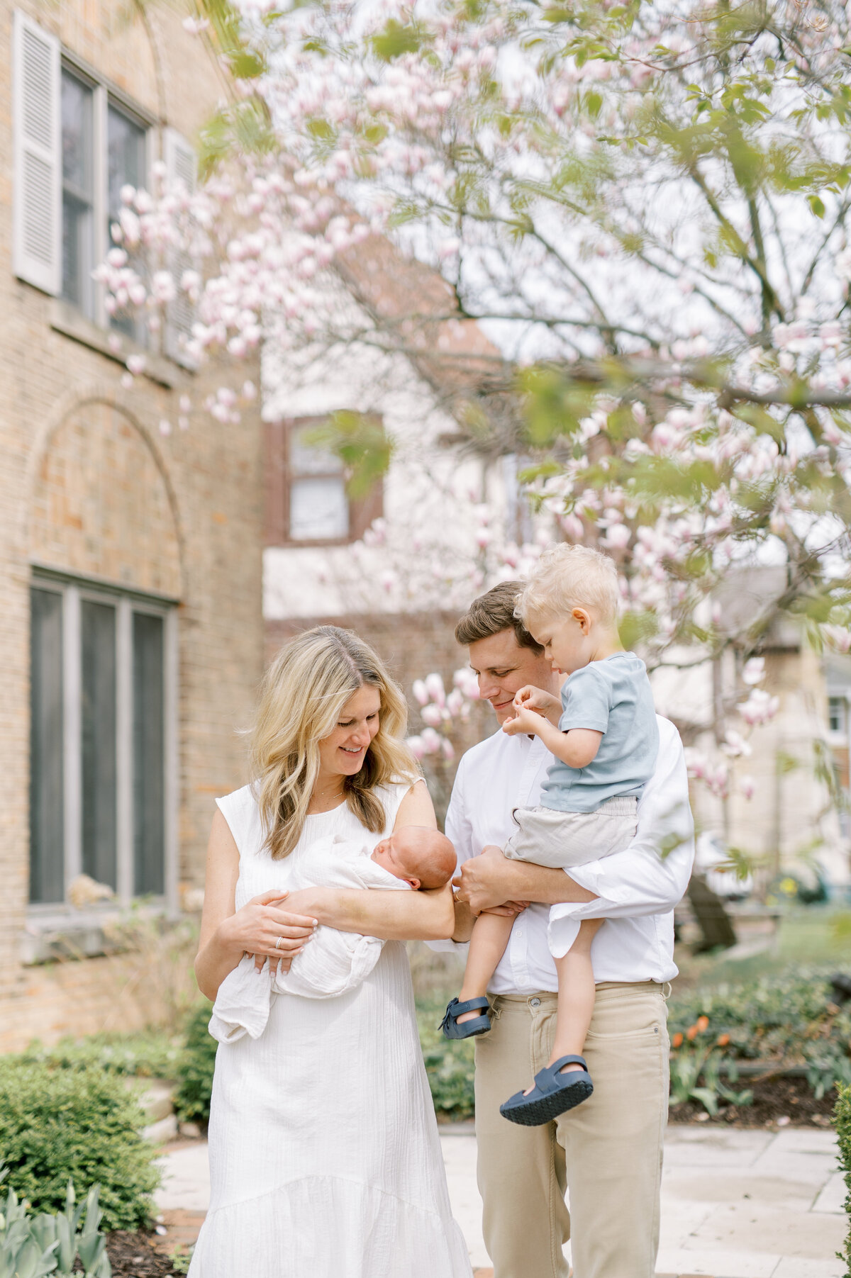 Outdoor family photo taken in Shorewood, Wisconsin of a family of four. Dad is holding the toddler and mom holds the baby during a photo session. Spring blooms are in the background