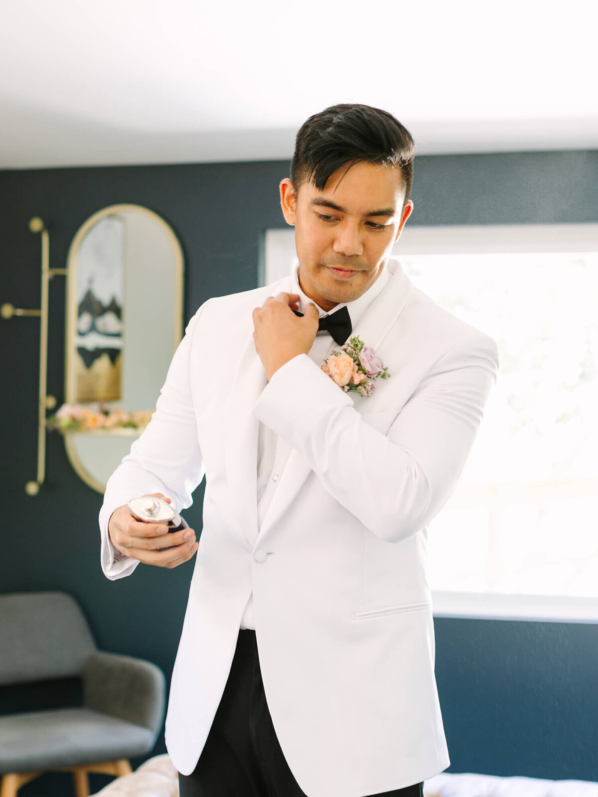 Groom in white tuxedo adjusting bow tie, holding a cologne spray. He wears a cream and purple floral boutonniere, standing in a bright room with modern decor.