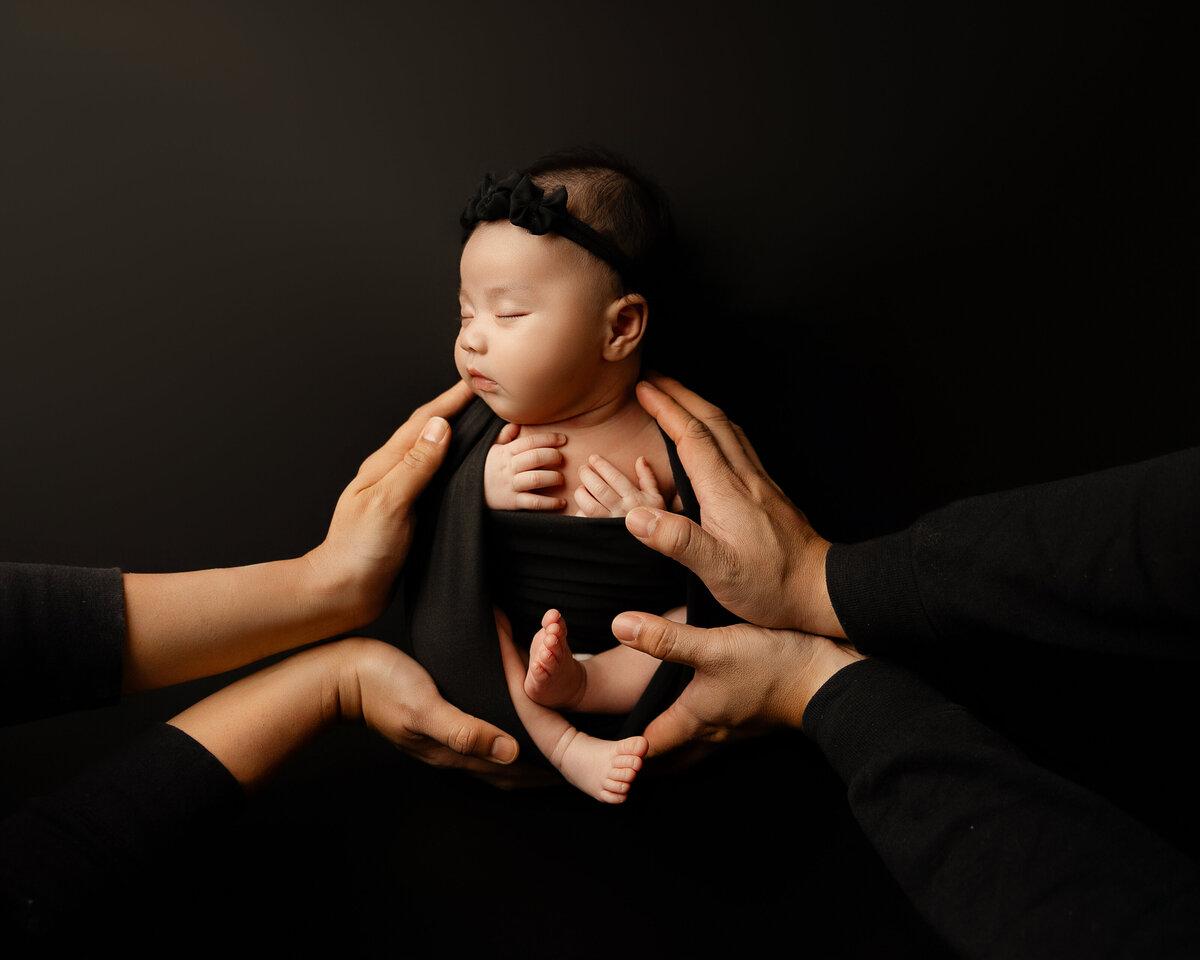 Fine art newborn session featuring a baby girl in a minimalist black setup, her parents’ hands creating a circle of love and connection around her. Captured by Inland Empire photographer Kaitlyn Dawn.