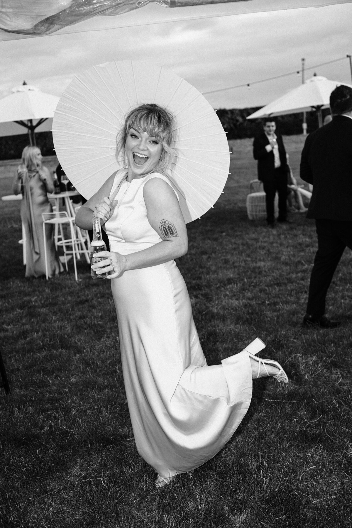 editorial style flash photo of wedding guest in satin dress holding a white parasol in front of umbrellas and outdoor furniture 