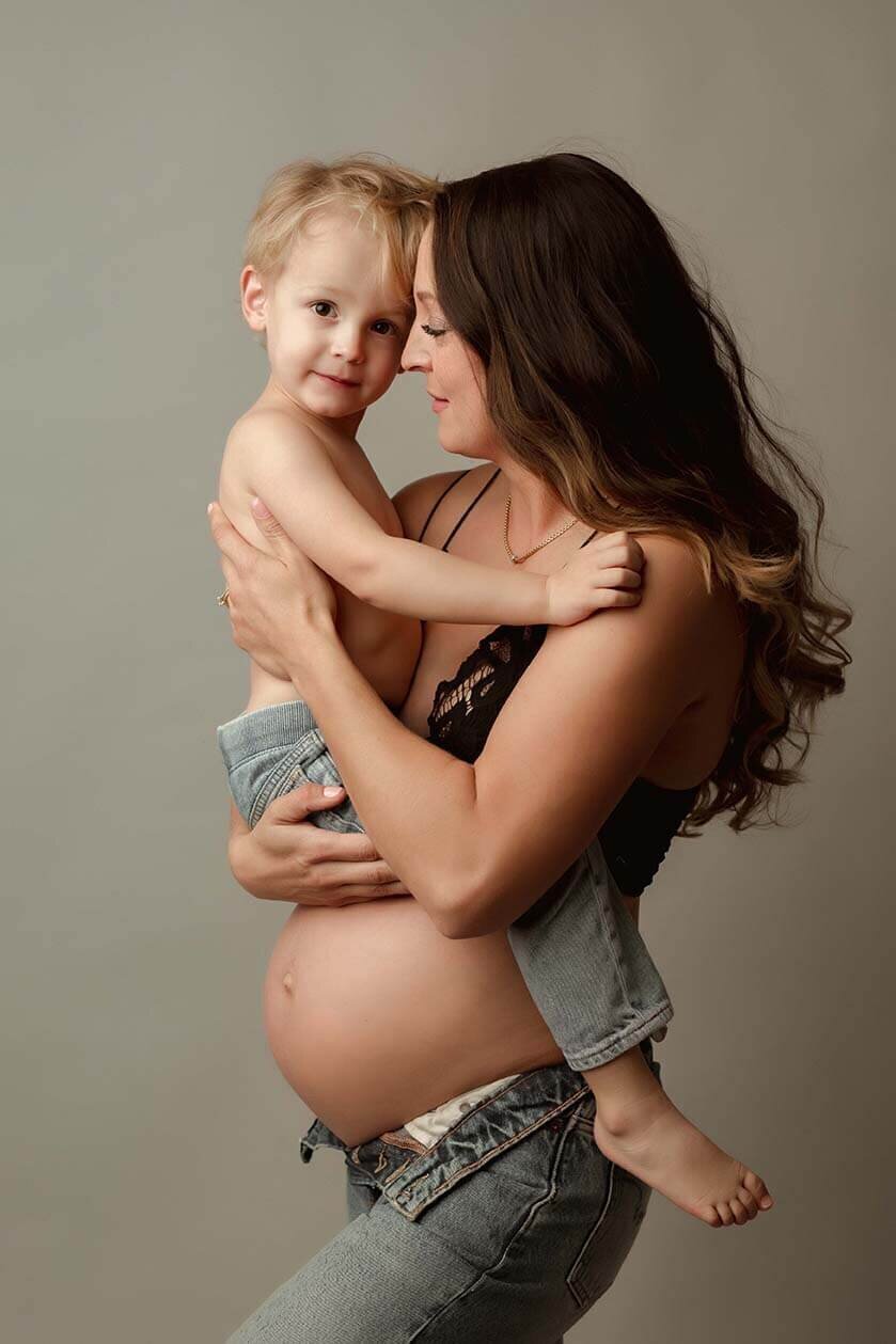 A mother holds her son over her pregnant belly. They are head to head but the boy is looking at the camera