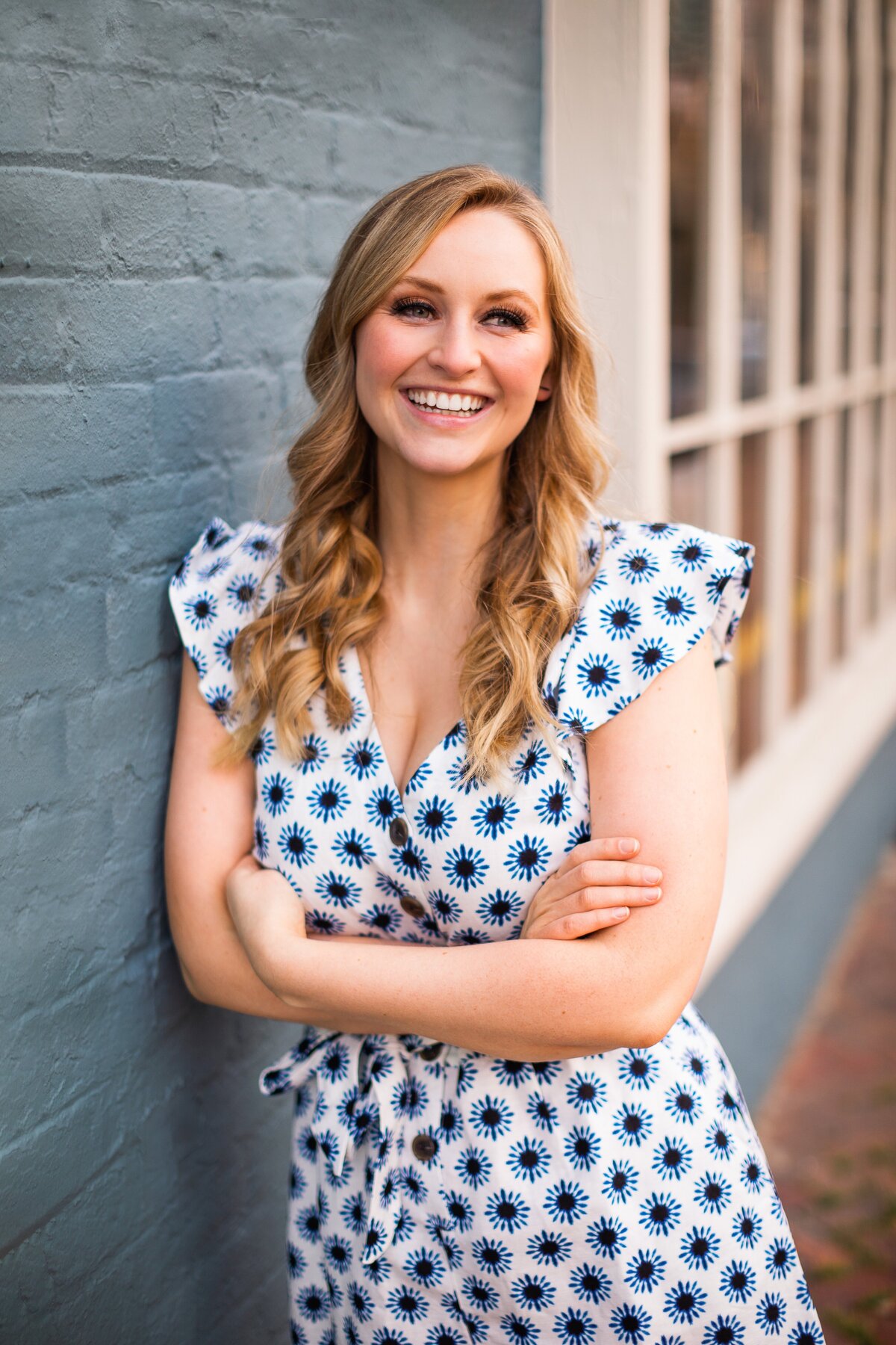 A woman with blonde curled hair wears a floral blue and white wrap dress while standing in front of a blue brick wall