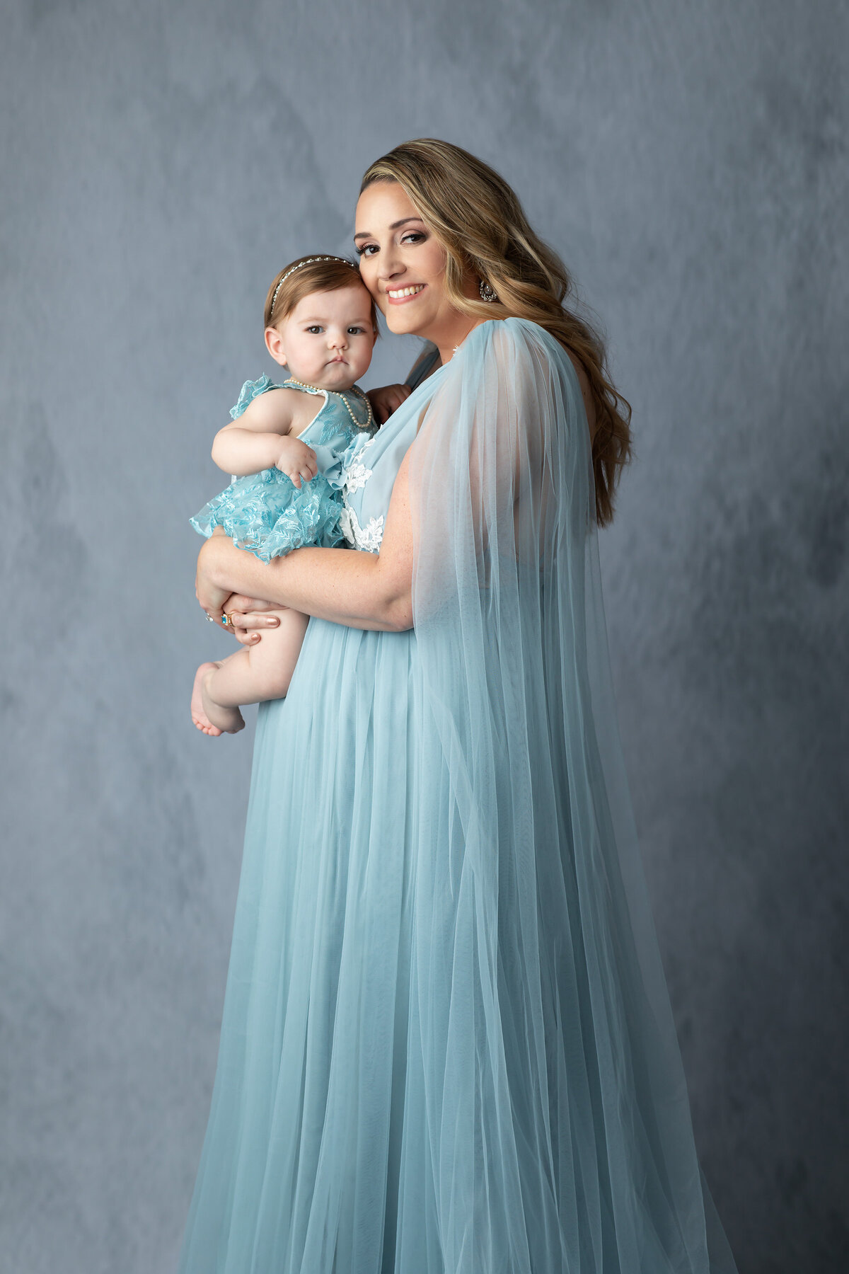 Mother and baby dressed in matching soft blue gowns share a loving moment during a Dallas milestone photographer session.