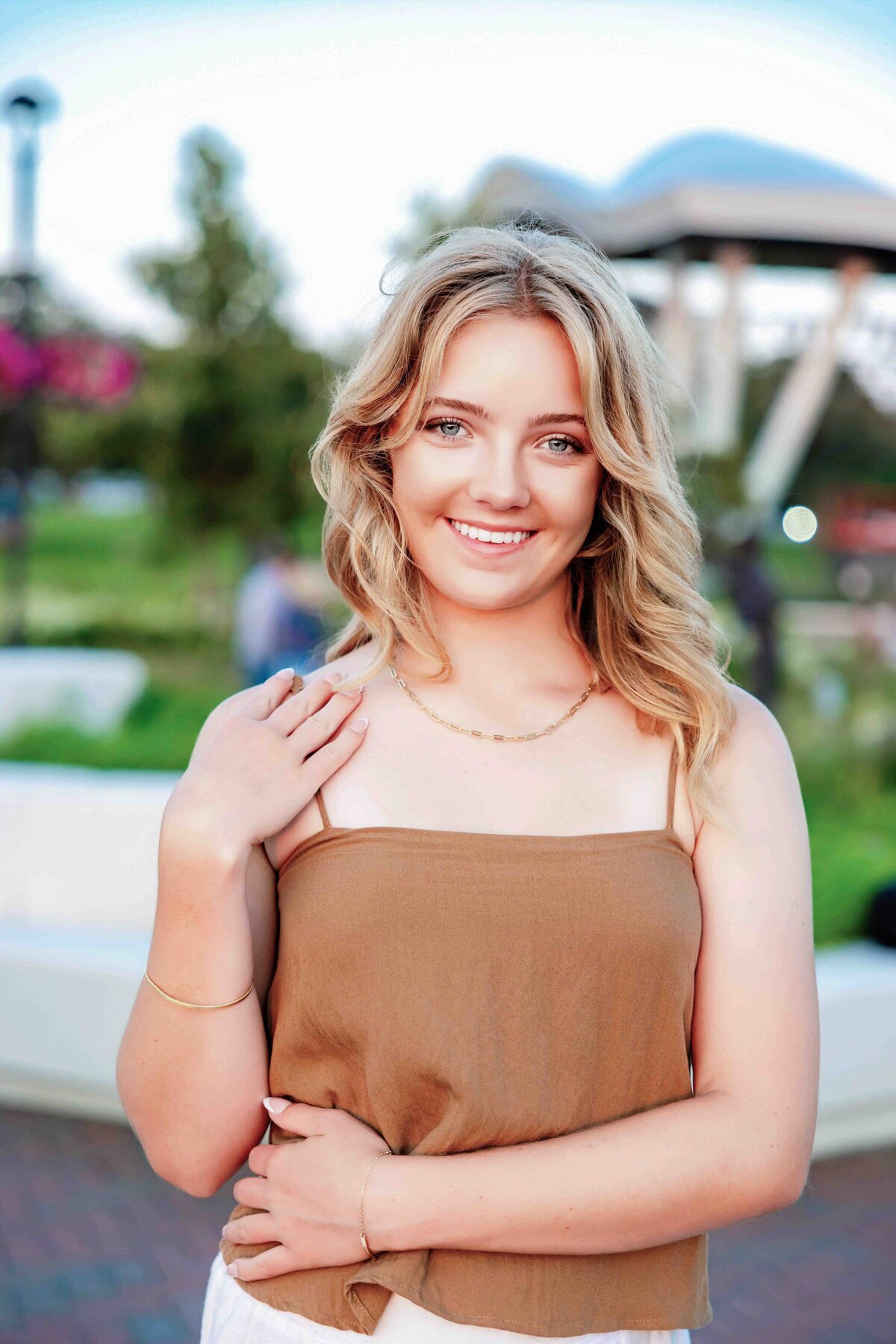 Outdoor senior portrait of a blonde girl in a tan top, softly smiling with modern architecture in the background.