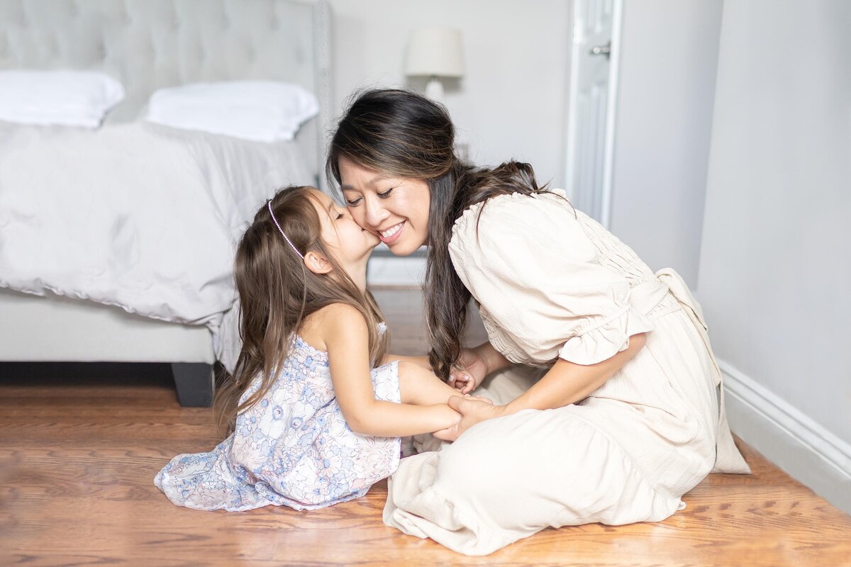 Photograph of a Mother and Daughter sitting together on the floor in front of a bed, mother holds the daughters hands and smiles while her 4 year old kisses her cheek in Virginia Beach