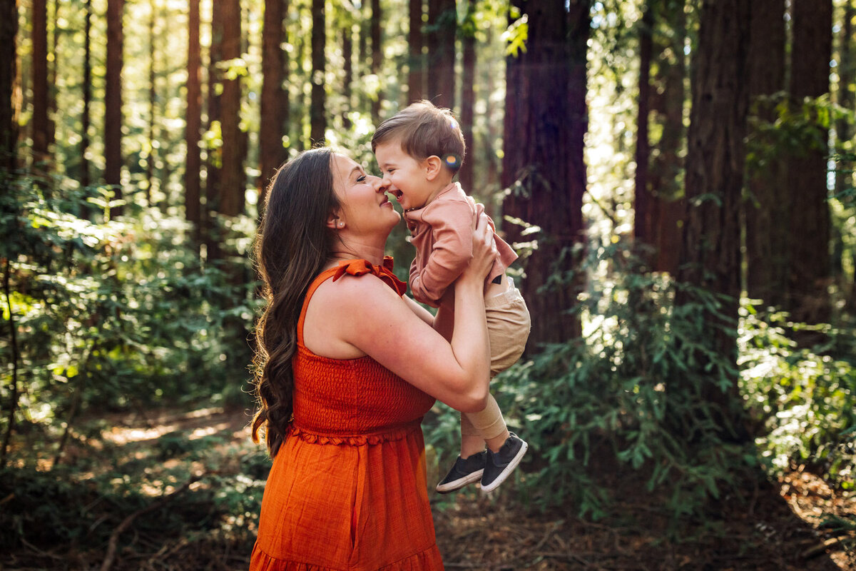 mom in orange dress in woods holding toddler son close to her space with them both smiling at each other